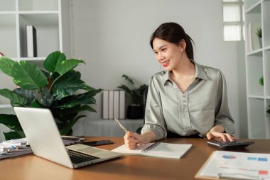 Evde çalışan genç bir kadın, dizüstü bilgisayar, defter ve hesap makinesi kullanarak modern bir ofis ortamında masa başında çalışıyor..