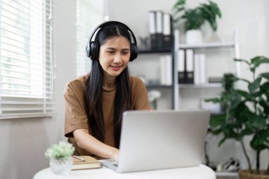 Kulaklık takan genç bir kadın, modern bir ofis ortamında dizüstü bilgisayarla çalışıyor, teknolojiye dayalı bir yaşam tarzı sergiliyor..