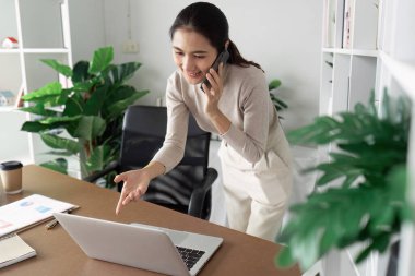 Genç profesyonel bir kadın ev ofisinde birden fazla iş yapıyor, telefonla konuşuyor ve dizüstü bilgisayar kullanıyor, arka planda yeşil bitkiler var..