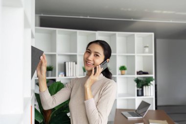 Neşeli genç bir kadın telefonda konuşarak ve şık, modern bir ofis ortamında kitapları düzenleyerek birden fazla iş yapıyor..