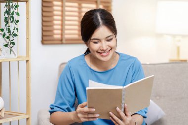 Gülümseyen genç bir kadın kitap okurken rahat, modern bir oturma odasında doğal ışık ve şık bir dekorla oturuyor..