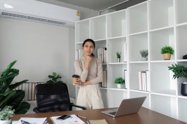 Çağdaş bir ofis ortamında elinde bir kahve fincanı, elinde bir dizüstü bilgisayar ve etrafında bitkiler olan profesyonel bir kadın..