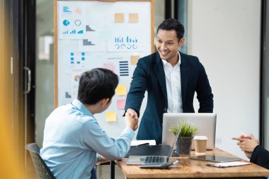 İki iş adamı modern ofis ortamında el sıkışıyor. Tablolar ve dizüstü bilgisayarlarla başarılı bir işbirliğini simgeliyor..