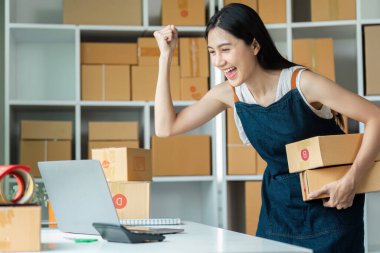Genç bir girişimci online iş başarısını kutluyor, paketleri tutuyor ve modern ofis ortamında bir dizüstü bilgisayar üzerinde çalışıyor..