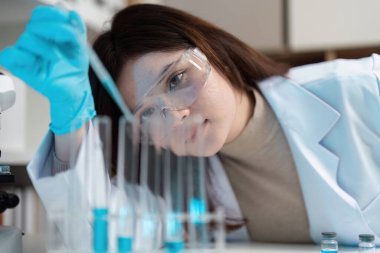 Modern bir laboratuvarda çalışan kadın bilim adamı test tüpleri kullanıyor ve koruyucu eldiven ve gözlük takıyor..