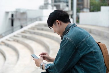 Dışarıda merdivenlerde oturan genç bir öğrenci, akıllı telefon kullanıyor. Günlük yaşam tarzı, modern teknoloji ve kampüsteki öğrenci hayatı..