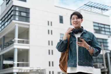 Sırt çantası ve kulaklığı olan genç bir öğrenci modern bir üniversite binasının önünde duruyor, akıllı telefon kullanıyor ve gülümsüyor..