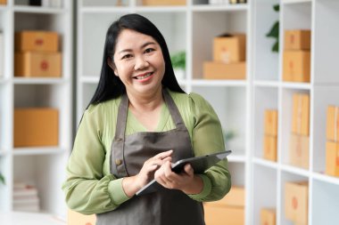 Evdeki ofiste paketlerle dolu raflarla çevrili bir tablet kullanan neşeli bir kadın online satış yapan bir şirketi temsil ediyor..