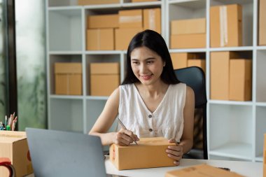 Küçük bir iş ofisinde çalışan genç bir kadın sevkiyat için paketler hazırlıyor. Arkaplanda kutularla dolu raflar.