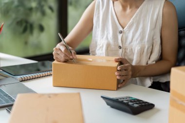 Küçük bir işletme sahibi, hesap makinesi ve defter ile birlikte bir kargo paketi üzerine yazı yazıyor..