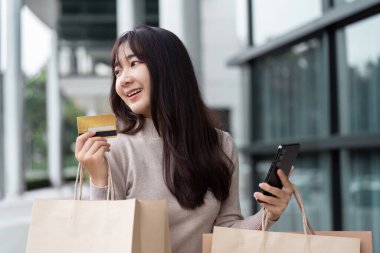 Alışveriş torbaları, kredi kartı ve akıllı telefonu olan neşeli bir kadın. Çağdaş bir perakende mağazasının önünde alışveriş yapmaktan zevk alıyor..