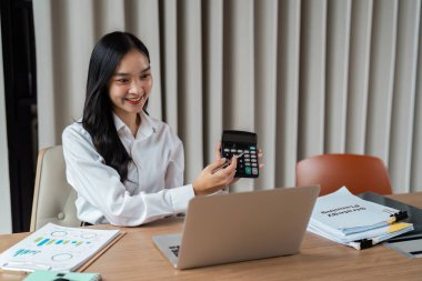 Günümüz ofis ortamında finansal belgeleri incelemek için hesap makinesi ve laptop kullanan kendine güvenen bir iş kadını..