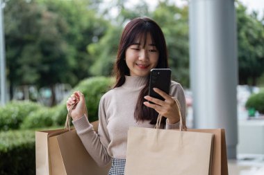 Alışveriş torbaları tutan ve Kara Cuma anlaşmalarını alışveriş merkezinin dışında akıllı telefonuyla kontrol eden genç bir kadın..