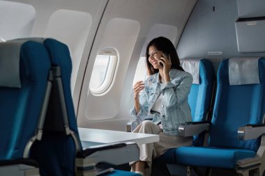 Kot ceketli genç bir kadın uçakta akıllı telefonunu kullanırken gülümsüyor ve pencereden manzaranın tadını çıkarıyor..