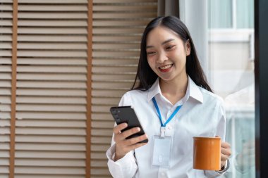 Akıllı telefon kullanan ve elinde kahve fincanı tutan kendine güvenen iş kadını, modern işyeri dinamiği ve dijital bağlantı örneğini veriyor.