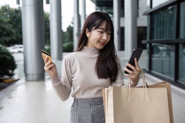 Alışveriş çantaları ve akıllı telefonlar taşıyan genç bir kadın, çağdaş bir mağazanın önünde Kara Cuma satışlarını kutluyor..