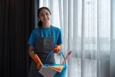 Gülümseyen temizlikçi elinde temizlik aletleriyle, çağdaş bir ev ortamında temizlik görevlerine hazır..