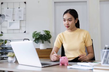 Genç bir kadın, bir dizüstü bilgisayar ve kumbara kullanarak, modern bir ofiste tasarruf ve akıllı para yönetimini sembolize ederek finansal planlamaya odaklanıyor..