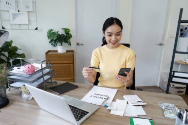 Genç bir kadın, ev ofis ortamında finansal planlama ve tasarruf için akıllı telefon ve kredi kartı kullanır.