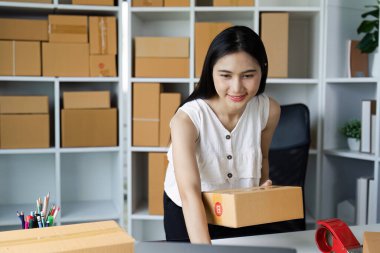 Gülümseyen kadın ev ofisinde kutu paketliyor, küçük işletme ve e-ticaret kavramlarını gözler önüne seriyor..