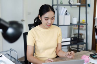 Genç bir kadın, dizüstü bilgisayar ve kredi kartı kullanarak para biriktirmeyi sembolize eden bir kumbarayla finansal planlamaya odaklanıyor..