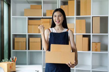 Önlüklü gülümseyen kadın, modern bir ofiste, paketlerle dolu raflarla çevrili, başarılı bir işi simgeleyen bir paket kutusu tutuyor..