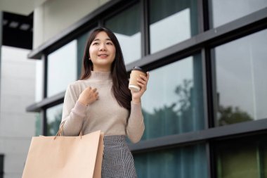 Çağdaş bir alışveriş merkezinin önünde çantalar ve kahveyle Kara Cuma alışverişi yapan genç bir kadın..