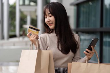 Alışveriş çantaları, kredi kartı ve akıllı telefonlu genç bir kadın, çağdaş bir alışveriş merkezinin önünde Kara Cuma gününün tadını çıkarıyor..