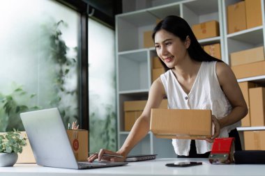 Çevrimiçi işinde çalışan gülümseyen bir kadın, elinde bir paket kutusu tutarken modern bir ev ofisinde dizüstü bilgisayar kullanıyor..