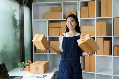 Modern bir ofiste karton kutular taşıyan gülümseyen genç bir kadın, e-ticaret ve online iş başarısını temsil ediyor..