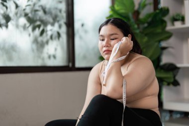 Düşünceli, kilolu bir kadın elinde bir mezurayla oturur, modern bir ev ortamında sağlık ve yaşam tarzını düşünür..