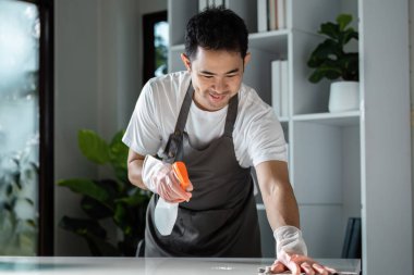 Önlük ve eldiven giyen bir adam parlak, modern bir oturma odasında bir sprey şişesiyle masayı temizliyor..