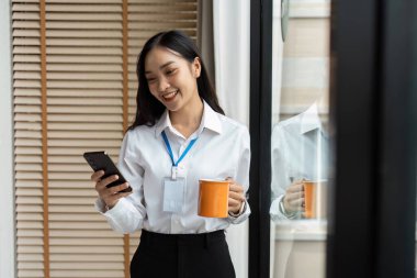 İş kadını moladan zevk alıyor, akıllı telefonunu kullanıyor ve elinde parlak, modern bir ofis ortamında kahve fincanı tutuyor..