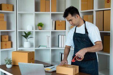 Önlüklü bir adam, raflar ve malzemelerle çevrili bir ofis ortamında nakliye için karton kutular paketliyor..