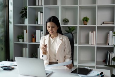 Modern ofis ortamında kendine güveni tam bir iş kadını. Etrafı kitaplarla ve bitkilerle çevrili bir dizüstü bilgisayarla çalışıyor..