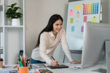 Grafik tasarımcısı, renkli tasarım elemanları ve araçlarıyla çevrili modern bir ofiste dijital tablet kullanır..