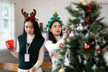 Two young women in festive attire decorate a Christmas tree, creating a joyful holiday atmosphere in a bright living room. clipart