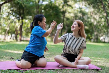 Parkta yoga minderinde oturan iki kadın, beşlik çakıyor ve açık havada güneşli bir günün tadını çıkarıyor..
