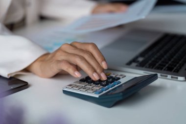 Bilgisayarı ve belgeleri olan bir masanın üzerinde hesap makinesi kullanarak finansal analizi ve iş planlamasını sembolize eden yakın plan bir el..