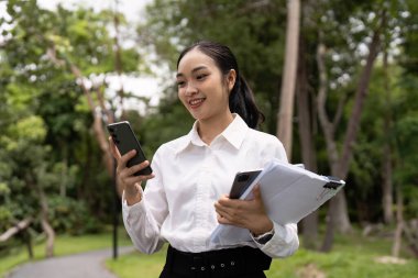 Beyaz tişörtlü profesyonel bir kadın elinde akıllı telefon ve belgelerle dışarıda iş yapıyor..