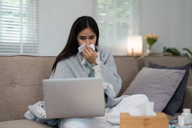 Kanepede dizüstü bilgisayar kullanarak battaniyeye sarılmış bir kadın, sıcak bir oturma odasında grip ya da grip belirtileri gösteriyor..