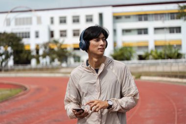 Pistte koşan genç bir adam, kulaklık ve açık renk ceket takıyor, kentsel bir ortamda spor ve sağlık koşullarını destekliyor..