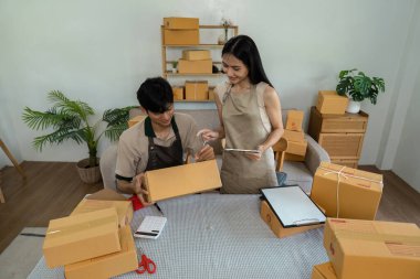 İki genç girişimci paketleri düzenliyor ve dijital cihazları kullanarak online mağazalarını modern bir çalışma alanında yönetiyorlar..