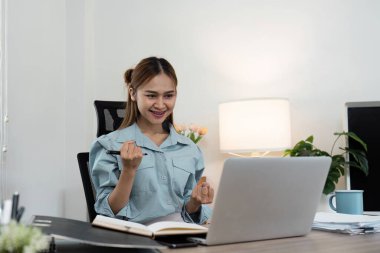 Evdeki genç bir iş kadını, belgeleri ve ofis dekoruyla çevrili dizüstü bilgisayarıyla çalışırken başarısını kutluyor..