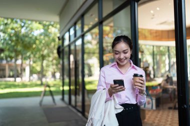 Pembe gömlekli bir iş kadını elinde kahve ve akıllı telefonla modern bir ofis binasının önünde mola veriyor..