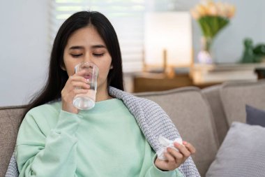 Genç bir kadın kanepeye oturur, battaniyeye sarınır, soğuk algınlığını veya grip belirtilerini yatıştırmak için su içer..