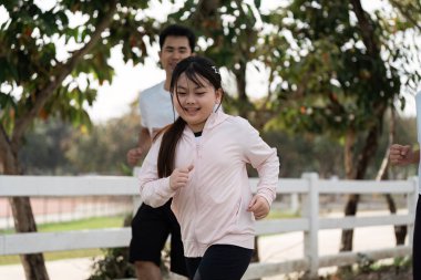 Parkta birlikte koşan bir aile güneşli bir günde arka planda beyaz bir çit olan açık havada egzersiz yapıyor..