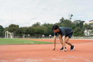 Odaklanmış bir atlet, canlı bir spor pistinde koşmaya kararlı ve hazır olduğunu gösteren başlangıç çizgisine çömelir..