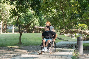 Tekerlekli sandalyedeki genç bir adam yemyeşil ve güneş ışığıyla çevrili bir arkadaşıyla parkta günün tadını çıkarıyor..