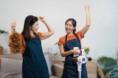 Bir evi temizleyen iki neşeli hizmetçi, rahat bir yaşam alanında takım çalışması ve modern temizlik aletleri sergiliyor..